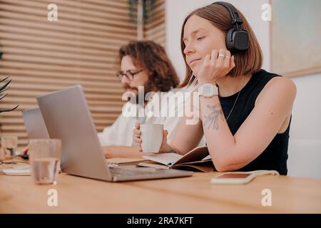 Donna freelance in cuffie che lavora su un computer portatile di sottofondo di un collega che lavora in coworking Foto Stock