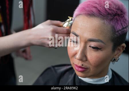 Il parrucchiere rasa il tempio di una cliente femminile. Donna asiatica con capelli corti rosa in barbiere. Foto Stock