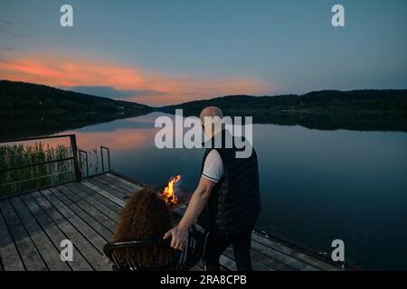 Coppia simpatica guarda il tramonto seduto accanto al fuoco sul molo, vista sul retro Foto Stock