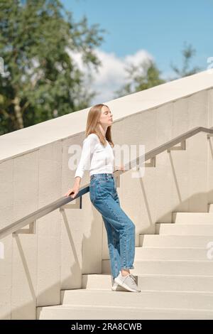 Donna carina in piedi con la schiena contro la ringhiera sulle scale e di fronte al sole Foto Stock