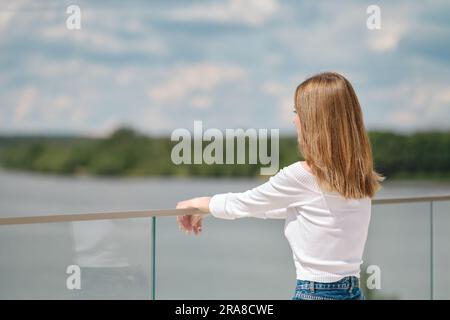 Donna irriconoscibile appoggiata su ringhiere di vetro sulla piattaforma di osservazione e guarda l'acqua Foto Stock