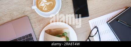 Colazione. Tazza di caffè cappuccino, sandwich, computer portatile e smartphone su un tavolo di legno nel caffè, vista dall'alto Foto Stock