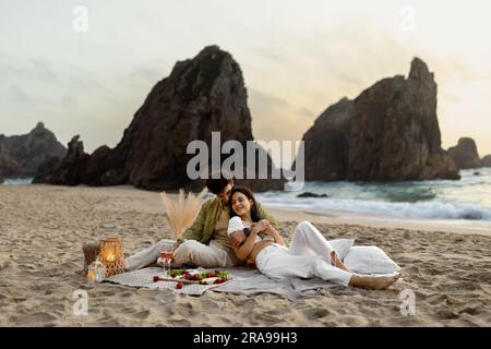 Coppie giovani che fanno un picnic romantico sulla spiaggia, abbracciano e si sdraiano su una coperta, celebrano il fidanzamento o l'anniversario Foto Stock