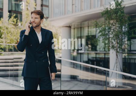 Un giovane lavoratore sicuro di sé che indossa una tuta formale cammina fuori, parla al telefono, tiene il portatile, si affretta a lavorare la mattina di sole. Foto Stock