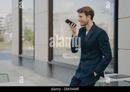 Un uomo d'affari sicuro di sé organizza la giornata, registra i messaggi, parla con il vivavoce, appoggiandosi al tavolo del caffè Foto Stock