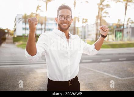 Allegro uomo d'affari europeo che alza le mani e rallegra la vittoria all'aperto per strada, sorridendo davanti alla telecamera Foto Stock