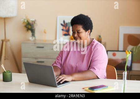 Donna afro-americana matura e felice con una t-shirt rosa seduta accanto alla scrivania di fronte al laptop in salotto, digitando sulla tastiera e collegando in rete Foto Stock