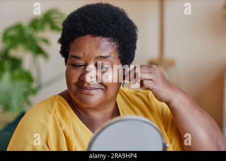 Donna matura sorridente che prova piccoli e graziosi orecchini di perle mentre si trova di fronte alla fotocamera e guarda allo specchio durante la preparazione dell'appuntamento Foto Stock