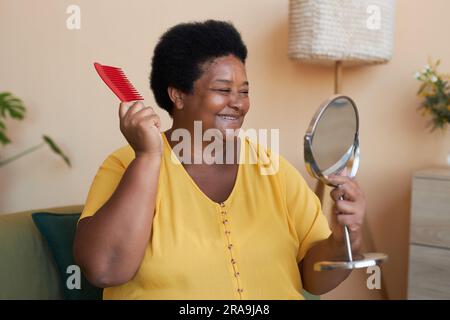 Allegra donna afroamericana matura con spazzola di plastica rossa che guarda allo specchio mentre si prende cura dei suoi capelli la mattina Foto Stock