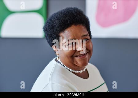 Felice donna afro-americana matura che guarda la macchina fotografica con un sorriso mentre si trova in uno studio d'arte contro opere d'arte appese a una parete grigia Foto Stock