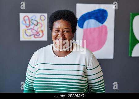 Felice insegnante d'arte maturo in abbigliamento casual davanti alla macchina fotografica contro opere d'arte appese a una parete grigia dello studio e che ti guarda Foto Stock