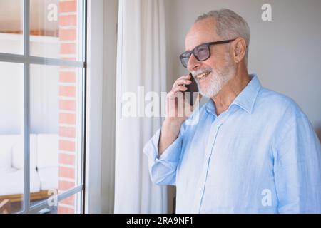 Allegro senior nonno parlando con il telefono vicino alla finestra a casa Foto Stock