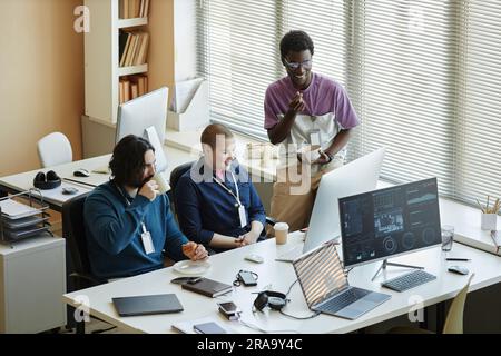 Tre giovani responsabili IT interculturali che si occupano di spuntini e guardano video o film online mentre sono seduti e in piedi sul posto di lavoro durante le pause Foto Stock