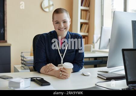 Giovane e grazioso responsabile DEL supporto IT con una tazza di caffè in mano seduto sul posto di lavoro di fronte al monitor del computer e guardato con sorriso Foto Stock