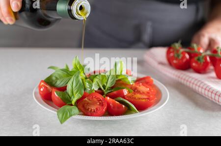 Versare l'olio d'oliva in bottiglia su un'insalata di pomodoro ciliegia con foglie di basilico Foto Stock