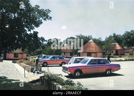 Auto moderne parcheggiate accanto a vecchie capanne di paglia, Kruger, Sud Africa. Data: Anni '1960 Foto Stock