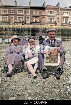 Tre vacanzieri, due donne e un uomo, seduti sulla spiaggia di ciottoli di Hastings, Sussex. Sta leggendo il quotidiano Daily Sketch. Sullo sfondo si può vedere una terrazza di hotel. Data: Giu-27 Foto Stock