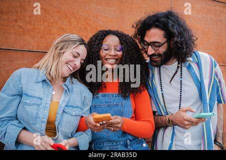 Donna gioiosa che usa il suo telefono cellulare mostrando ai suoi amici divertenti messaggi di testo e divertendosi insieme. Gruppo sorridente di amici che si divertono a navigare su Internet con lo smartphone, giovani con il cellulare. Foto di alta qualità Foto Stock