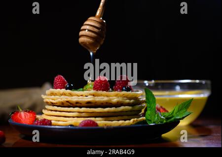 Bastoncino di legno a forma di favo con miele che gocciola su pancake appena sfornati con frutti di bosco selvatici per Shrovetide Foto Stock