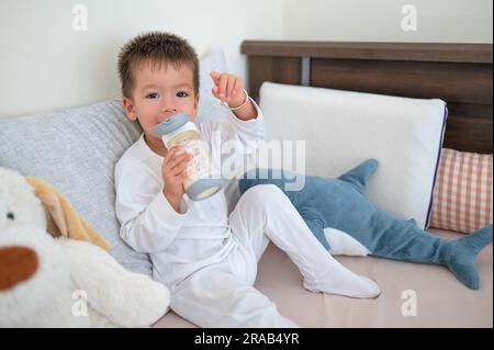 il bambino di 2 anni si siede sul letto e beve il latte da un biberon dotato di paglia, circondato da amici giocattolo al mattino che indossano un pigiama Foto Stock