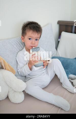 il bambino di 2 anni si siede sul letto e beve il latte da un biberon dotato di paglia, circondato da amici giocattolo al mattino che indossano un pigiama Foto Stock
