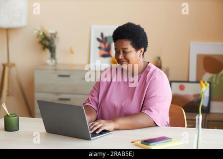 Donna afro-americana matura e felice con una t-shirt rosa seduta accanto alla scrivania di fronte al laptop in salotto, digitando sulla tastiera e collegando in rete Foto Stock