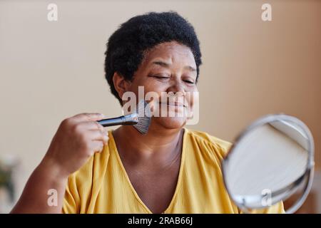 Donna afro-americana sorridente che tocca il suo viso con la spazzola mentre applica polvere sciolta sulla pelle e guarda allo specchio durante il trucco Foto Stock