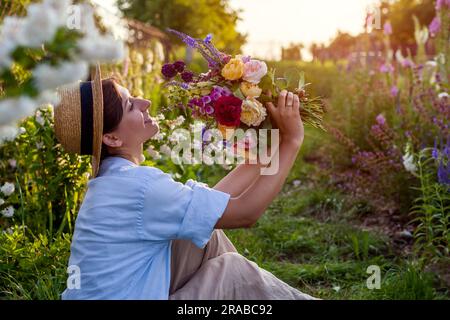 Donna giardiniera che si gode il giardino estivo al tramonto con rose fresche mescolate con veronika e guanti di volpe. Il contadino ha raccolto il bouquet di fiori e si è rilassato su g Foto Stock