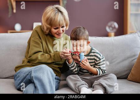 Ritratto frontale di madre e figlio con sindrome di Down che soffia bolle mentre si siede sul divano a casa Foto Stock