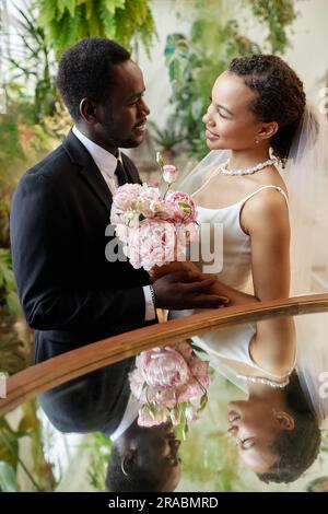 Ritratto verticale della giovane coppia nera che si sposa e si guarda a vicenda in orangerie con riflessi a specchio Foto Stock