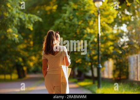 Ora legale. Vista da dietro donna alla moda con camicia bianca e borsa tote che cammina nel parco cittadino. Foto Stock