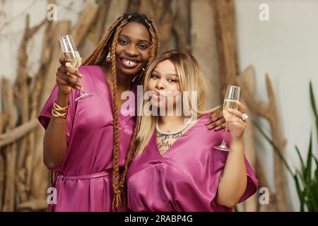 Indossa un ritratto in vita di due donne nere come damigelle d'onore che indossano una macchina fotografica rosa durante il ricevimento di nozze e tengono in mano dei bicchieri di champagne Foto Stock