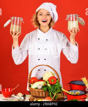 Lo chef sorridente in uniforme bianca in piedi al tavolo con cesto di verdure contiene vasetti di vetro con semole. Cucina sana. La cuoca femmina tiene i vasetti con Foto Stock