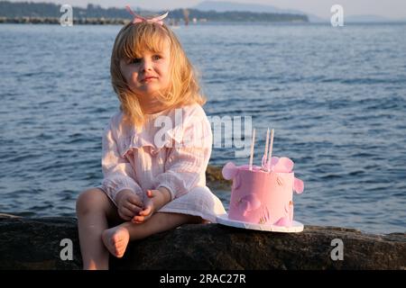 piccola ragazza triste seduta che si gratta la gamba sulla riva di un lago marino con una torta. in solitaria occasione del terzo anniversario della sessione fotografica, vestito da fiocco rosa stanco, capelli biondi biondi Foto Stock