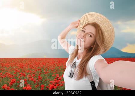 Bella donna in cappello di paglia che scatta selfie in fiori di papavero vicino alla montagna Foto Stock