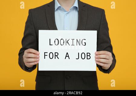 Uomo disoccupato con un cartello con una frase in cerca di lavoro su sfondo arancione, primo piano Foto Stock