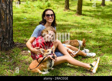 La madre abbraccia la sua piccola figlia riposa sulla sua vecchia con gli occhi chiusi, con due cani corgi contro l'erba verde nella foresta Foto Stock