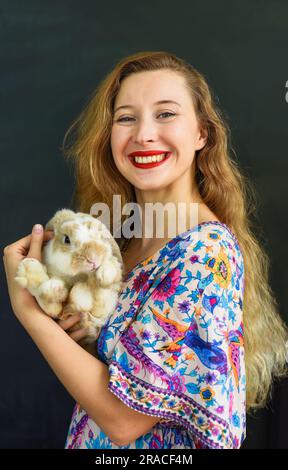 Bella donna russa con lunghi capelli biondi che tiene in mano un animale domestico di coniglio contro il muro nero Foto Stock