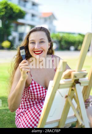 Giovane artista che tiene in mano un barattolo con pennelli e cavalletto vicino al mare e alle palme in estate Foto Stock