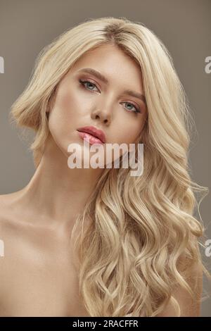 Scatto verticale di un ritratto di una bella donna con capelli biondi ricci e pelle pulita, foto di bellezza da studio su sfondo beige Foto Stock