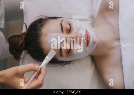 Vista dall'alto di una femmina anonima che applica una maschera facciale sul viso mentre si sdraia sul letto con gli occhi chiusi Foto Stock