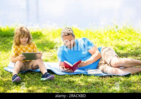Il padre forma l'educazione del figlio con un libro e un tablet. Padre e figlio si rilassano nel parco. educazione scolastica dell'infanzia. figlio con padre che si unisce in estate. famiglia Foto Stock