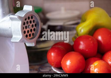 tritacarne elettrico con pepe sul tavolo accanto ai pomodori e carote sul tavolo in cucina, cucina adijika fatta in casa, preparatio fatto in casa Foto Stock