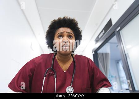 Medico donna afroamericana che indossa scrub, che corre attraverso il corridoio all'ospedale. Ospedale, medicina, sanità e lavoro, inalterati. Foto Stock