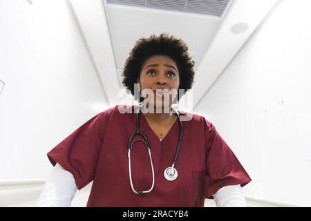 Medico donna afroamericana che indossa scrub, che corre attraverso il corridoio all'ospedale. Ospedale, medicina, sanità e lavoro, inalterati. Foto Stock