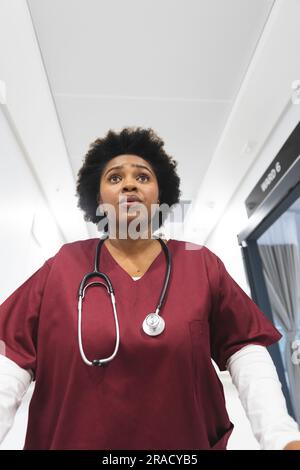 Medico donna afroamericana che indossa scrub, che corre attraverso il corridoio all'ospedale. Ospedale, medicina, sanità e lavoro, inalterati. Foto Stock