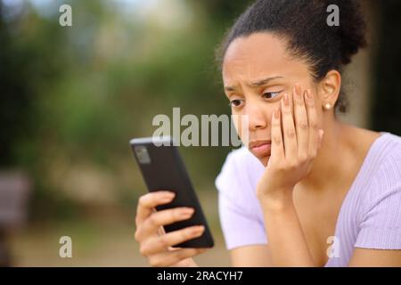 Preoccupata donna nera che controlla il cellulare seduto in un parco Foto Stock