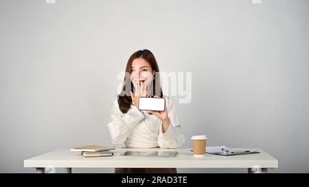 Una giovane donna d'affari asiatica sorpresa e allegra siede alla sua scrivania, coprendo la bocca con un'espressione facciale scioccata e tenendo in mano lo smartphone Foto Stock