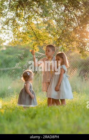 tre bambini piccoli che soffiano bolle di sapone in estate al tramonto in un parco vicino a un albero, spazio per il testo. Foto di alta qualità Foto Stock