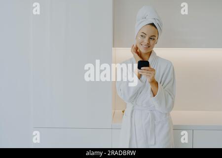 Una donna stupenda si prende cura della carnagione, applica la crema, sorride delicatamente, si posa con cura in una veste bianca a casa. Foto Stock